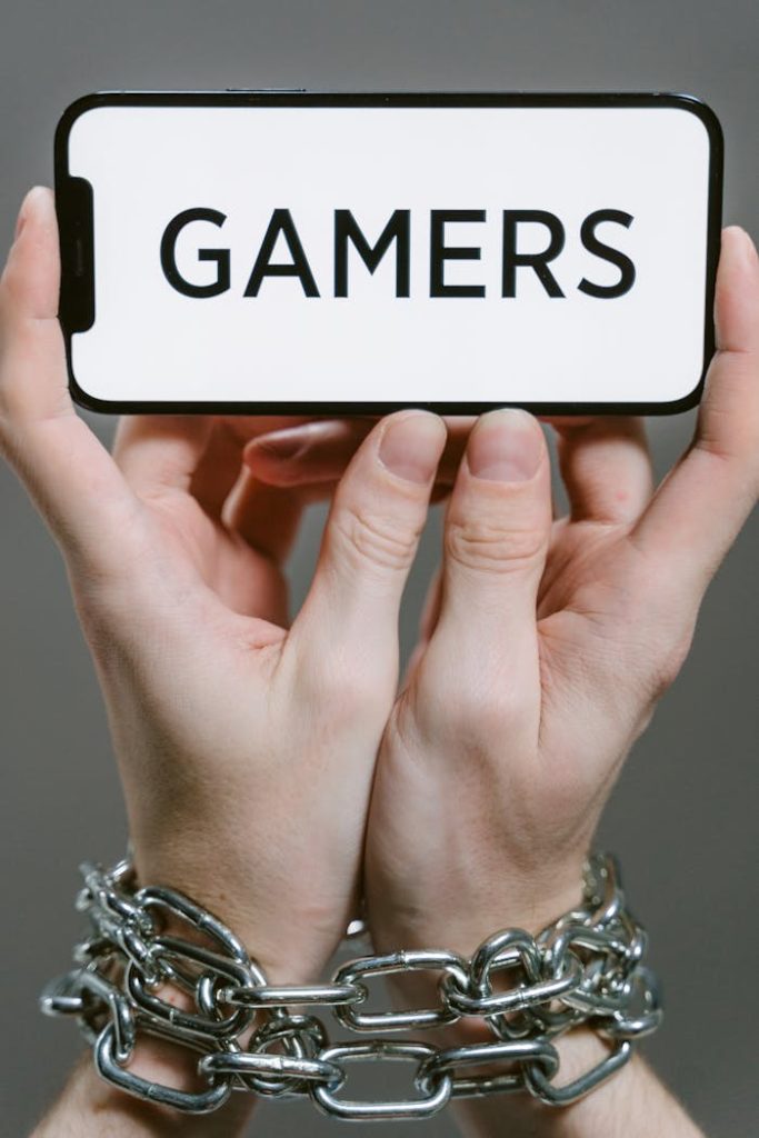 Close-up of hands bound by chains holding a phone displaying Gamers, symbolizing gaming addiction.